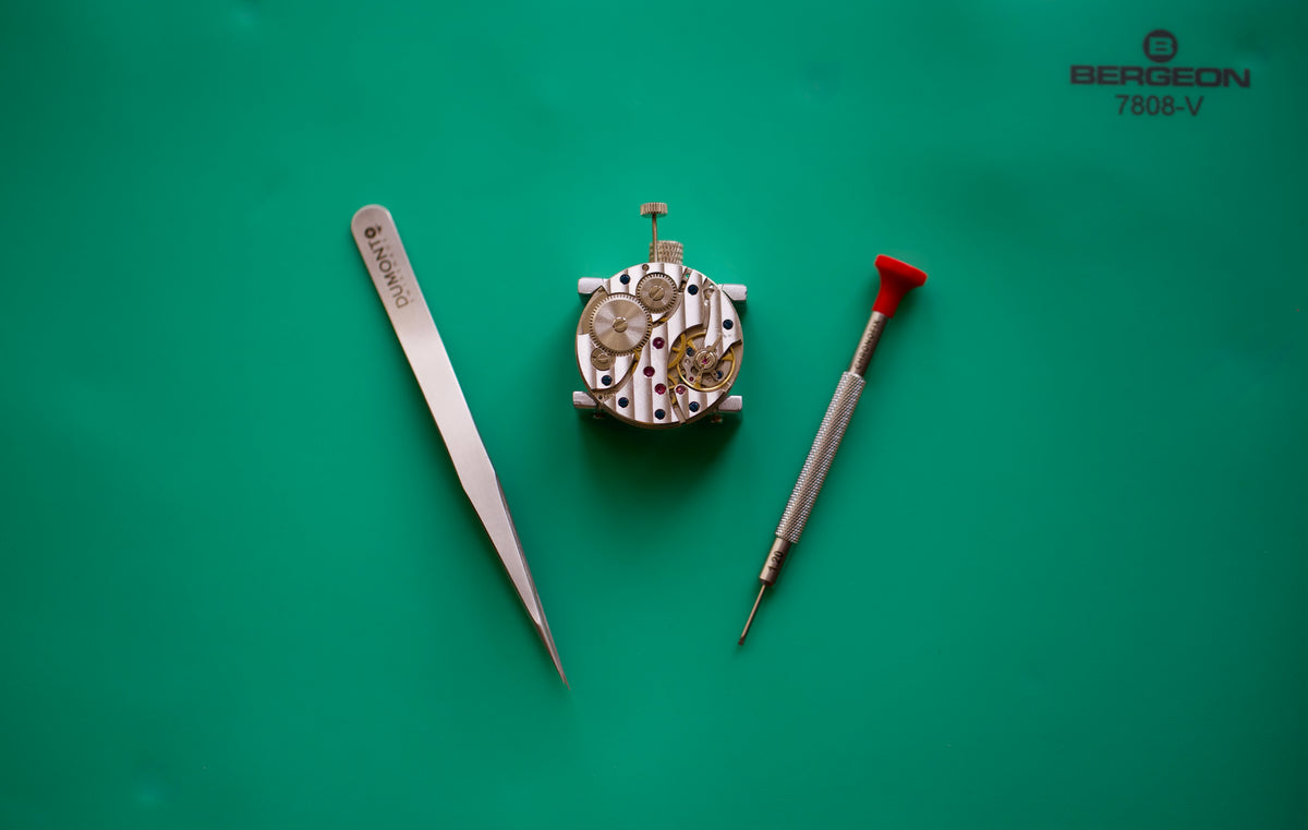 A watch movement mechanism displayed on a solid green background, accompanied by a silver dumont tweezers tool and Chronofix screwdriver with a red handle. The Bergdéon 7808-V logo is visible in the top right corner.